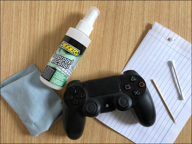 A DualShock 4 Controller sitting on top of a piece of a paper next to a toothpick, a Q-tip, a spray bottle of isopropyl alcohol, and a cloth. 